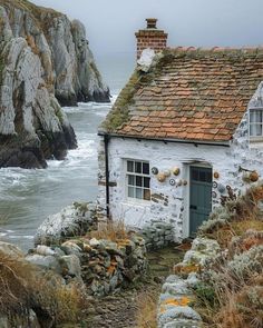 an old white house sitting on the side of a cliff next to the ocean with waves crashing in front of it