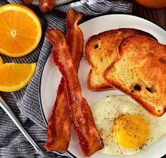 an egg, bacon and toast on a white plate with oranges next to it