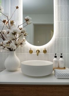 a white sink sitting under a mirror next to a vase filled with flowers and soap