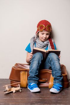 a little boy sitting on top of a piece of luggage reading a book with goggles