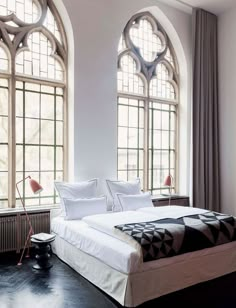 a large bed sitting next to two tall windows in a white room with black flooring