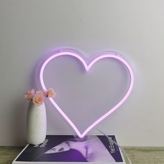 a pink heart shaped neon sign sitting on top of a table next to a vase with flowers