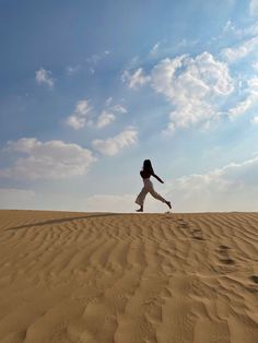 a woman standing on top of a sandy dune under a blue sky with white clouds