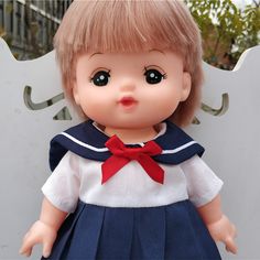 a doll with blonde hair wearing a blue dress and red bow around her neck is standing in front of a white fence