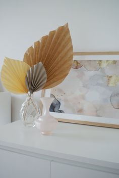 a white dresser topped with a vase filled with flowers next to a painting and fan