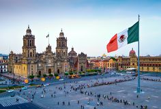 the mexican flag is flying in front of an old building with many people walking around it