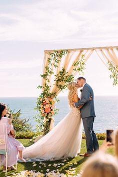 a bride and groom kissing under an arch with flowers on the grass by the ocean