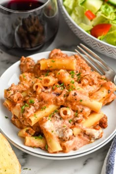 pasta with meat and sauce on a white plate next to a bowl of salad, fork and glass of red wine