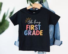 a t - shirt that says oh hey first grade hanging on a clothes rack next to a potted plant