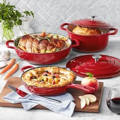 three red casserole dishes on a marble countertop with vegetables and wine glasses