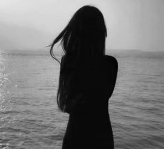 a woman standing in front of the ocean with her back to the camera, looking at the water