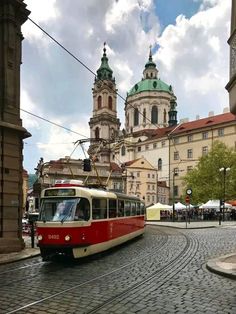 a red and white train traveling down a street next to tall buildings with green tops