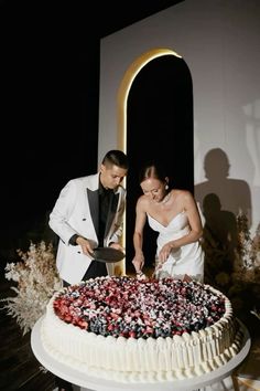 a man and woman cutting into a cake with berries on it in front of a black background