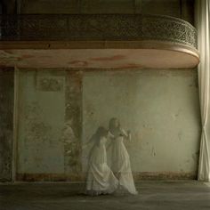 two women in white dresses standing next to each other near a window with drapes on it