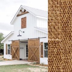 a white barn with wooden doors and windows next to a tall grass mat on the ground