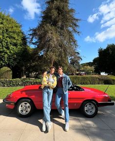 two people standing next to a red sports car in front of a tree and bushes