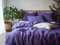 a bed with purple sheets and pillows in a room next to a potted plant