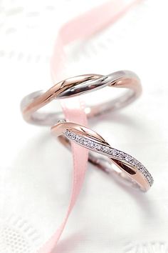 two wedding rings with diamonds on them sitting on a white tablecloth next to a pink ribbon