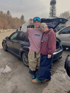 a man and woman standing in front of a car with the hood up, posing for a photo