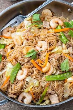 stir fried noodles with shrimp and vegetables in a skillet on a wooden table, ready to be eaten