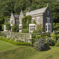 a large stone house surrounded by greenery and flowers in the foreground, with trees behind it