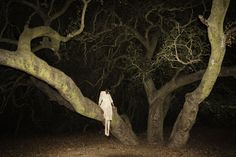 black and white photograph of person standing in front of trees at night with head on tree branch