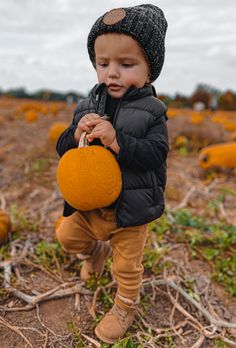 a little boy in a black jacket and hat holding an orange pumpkin on a field