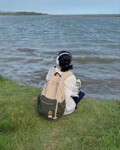 a person sitting on the grass near water with a backpack in front of them, looking at the water