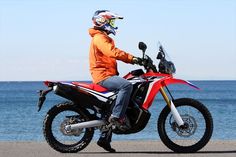 a man riding on the back of a red dirt bike next to the ocean in front of a blue sky