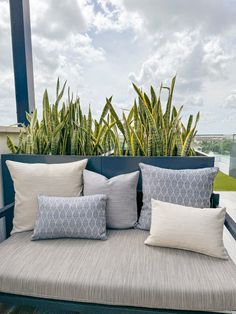 a couch with several pillows on it sitting in front of some plants and bushes outside