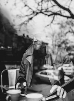 a man with a beard sitting at a table in front of a window looking into the distance