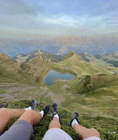 two people sitting on top of a grass covered hill with mountains in the background and lake below