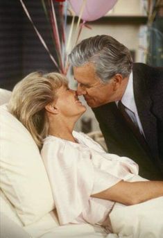 an older man and woman kissing on the bed with balloons in the air behind them