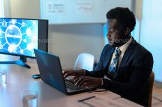 a man sitting in front of a laptop computer on top of a desk with a monitor behind him