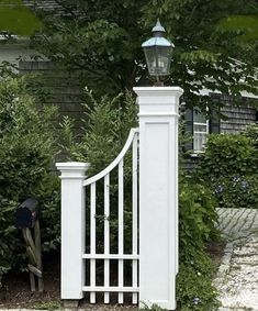 a white gate with a light on top in front of some bushes and trees near a house