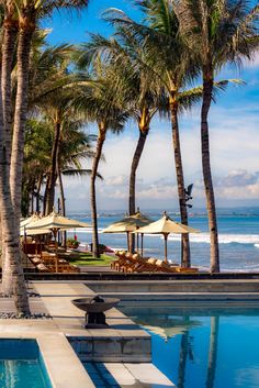 an outdoor swimming pool surrounded by palm trees and the ocean in the background with chairs under umbrellas