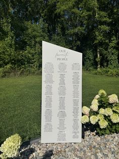 a large white sign sitting in the middle of a field next to flowers and trees