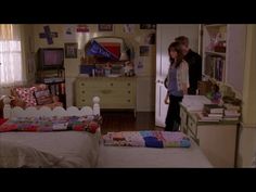 two people standing in a bedroom next to a bed and dresser with books on it