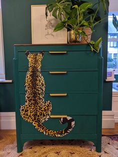 a green dresser with a leopard painted on the top and bottom drawer, next to a potted plant