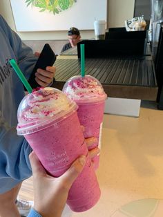 two people holding up pink drinks in front of a coffee shop counter, one is holding a cell phone