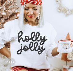 a woman wearing a merry sweatshirt and santa clause hat sitting in front of a christmas tree