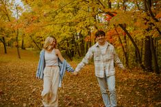 a man and woman holding hands while walking through the woods with fall leaves on the ground