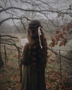 a woman with long hair standing in front of a fence holding an open book and looking into the distance