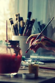 a person holding a paintbrush in front of a jar filled with paints and brushes