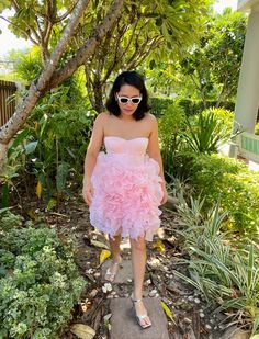 a woman in a pink dress and sunglasses standing on a sidewalk near some trees, bushes and plants