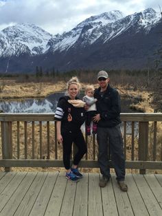 a man and woman standing on a deck with a baby in front of snowy mountains