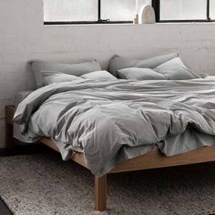 an unmade bed with grey sheets and pillows in front of two large window sills