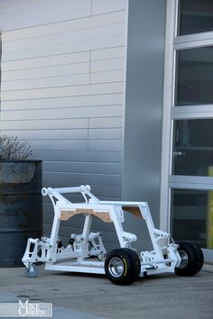 a small white vehicle with wheels parked in front of a gray building next to a potted plant