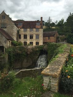 an old stone building with a waterfall in the foreground and flowers growing on the other side