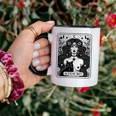 a woman's hand holding a white and black coffee mug with the witch tarot card on it